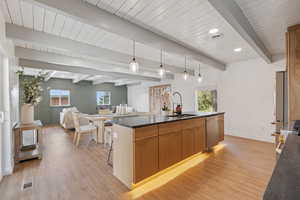 Kitchen with decorative light fixtures, a kitchen island with sink, dark countertops, beam ceiling, and light wood finished floors