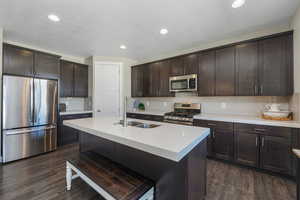 Kitchen featuring dark brown cabinets, stainless steel appliances, backsplash, a kitchen island with sink, and dark wood-style flooring