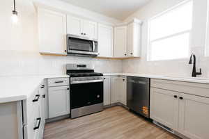 Kitchen with stainless steel appliances, light wood-style floors, decorative light fixtures, backsplash, and white cabinetry