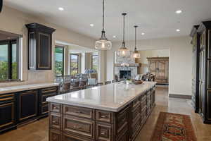Kitchen with dark brown cabinets, a stone fireplace, a kitchen island, light stone counters, and recessed lighting
