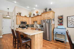 Kitchen with appliances with stainless steel finishes, light brown cabinets, a center island, light wood-type flooring, and decorative light fixtures