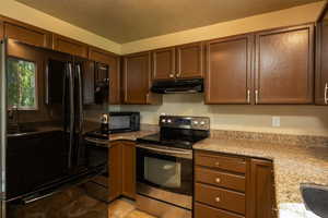 Kitchen featuring black appliances, under cabinet range hood, a textured ceiling, light stone counters, and brown cabinetry