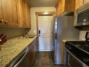 2nd Kitchen featuring appliances with stainless steel finishes, a textured ceiling, light stone countertops, brown cabinetry, and dark stone finish floors