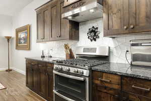 Kitchen with double oven range, dark brown cabinets, under cabinet range hood, backsplash, and dark stone countertops