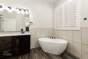 Full bath featuring vanity, a soaking tub, dark wood-style flooring, tile walls, and wainscoting