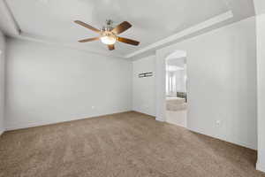 Empty room with a raised ceiling, light carpet, arched walkways, and ceiling fan