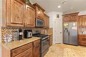 Kitchen with appliances with stainless steel finishes, tasteful backsplash, brown cabinetry, light stone counters, and stone tile flooring