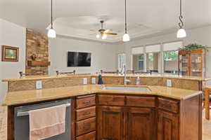 Kitchen with hanging light fixtures, a raised ceiling, brown cabinets, stainless steel dishwasher, and light stone countertops