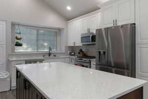 Kitchen featuring appliances with stainless steel finishes, light stone countertops, white cabinetry, decorative backsplash, and vaulted ceiling