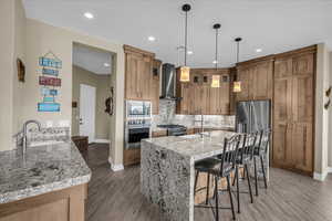 Kitchen featuring a kitchen bar, light stone countertops, stainless steel appliances, an island with sink, and wall chimney range hood