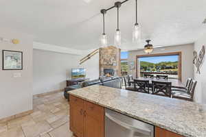 Kitchen with brown cabinetry, dishwasher, hanging light fixtures, light stone countertops, and a stone fireplace