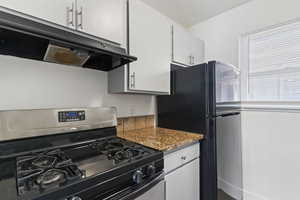 Kitchen with under cabinet range hood, stainless steel gas range, white cabinetry, and dark stone counters