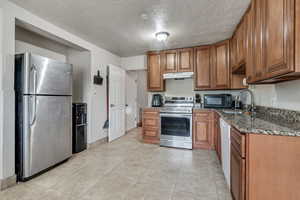 Kitchen featuring appliances with stainless steel finishes, dark stone counters, brown cabinets, a textured ceiling, and light tile patterned floors