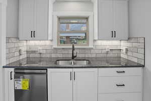 Kitchen with stainless steel dishwasher, dark stone countertops, white cabinetry, and tasteful backsplash