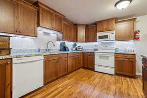 Kitchen featuring white appliances, brown cabinetry, a textured ceiling, light wood-type flooring, and light stone countertops