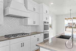 Kitchen with white cabinets, stainless steel appliances, decorative backsplash, custom exhaust hood, and decorative light fixtures