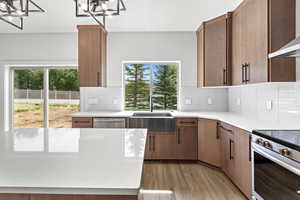 Kitchen with ventilation hood, appliances with stainless steel finishes, light stone counters, light wood-style floors, and brown cabinetry