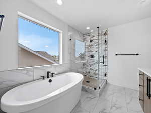 Bathroom featuring a freestanding bath, vanity, light marble finish floors, a marble finish shower, and tile walls