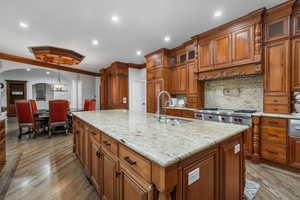 Kitchen featuring brown cabinetry, backsplash, light stone counters, glass insert cabinets, and ornamental molding