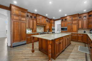Kitchen with glass insert cabinets, brown cabinetry, light stone counters, an island with sink, and backsplash