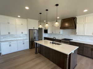 Kitchen featuring premium range hood, white cabinets, appliances with stainless steel finishes, a center island with sink, and pendant lighting