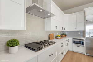 Kitchen featuring wall chimney exhaust hood, white cabinets, stainless steel appliances, decorative backsplash, and light stone countertops