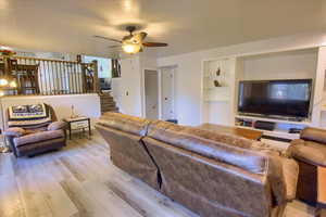 Living room featuring light wood-type flooring, ceiling fan, stairway, and built in shelves