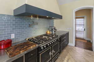 Kitchen featuring ventilation hood, decorative backsplash, high end black range oven, dark cabinets, and light tile patterned flooring