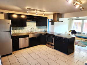 Kitchen with dark cabinets, appliances with stainless steel finishes, ventilation hood, backsplash, and track lighting