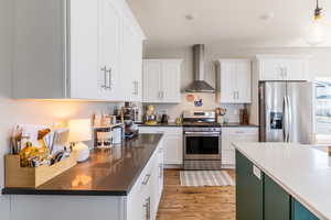 Kitchen with appliances with stainless steel finishes, wall chimney range hood, hanging light fixtures, light wood-style flooring, and white cabinetry