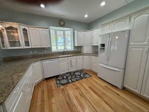 Kitchen with white appliances, glass insert cabinets, light wood-style flooring, dark stone counters, and white cabinetry