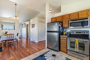 Kitchen featuring stainless steel appliances, brown cabinetry, lofted ceiling, light tile patterned floors, and a chandelier