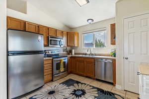 Kitchen featuring appliances with stainless steel finishes, brown cabinetry, vaulted ceiling, light tile patterned floors, and light countertops