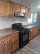 Kitchen with black range with gas stovetop, under cabinet range hood, dark wood-style floors, and brown cabinets