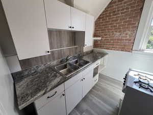 Kitchen featuring white cabinets, light wood finished floors, brick wall, dark countertops, and vaulted ceiling