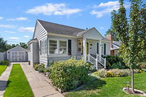 Bungalow with a front yard, a garage, a shed, a shingled roof, and driveway