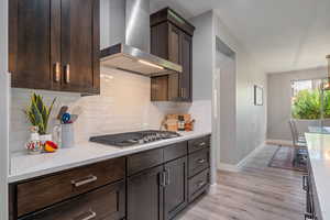 Kitchen with wall chimney range hood, dark brown cabinets, tasteful backsplash, light stone counters, and light wood finished floors