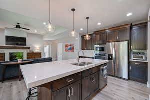 Kitchen featuring dark brown cabinets, appliances with stainless steel finishes, pendant lighting, open floor plan, quartz countertops, and a kitchen island with sink