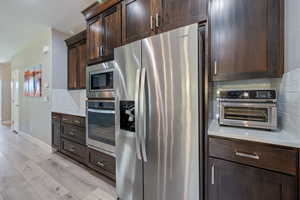 Kitchen with backsplash, appliances with stainless steel finishes, dark brown cabinets, and light wood-type flooring