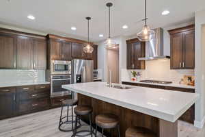 Kitchen featuring dark brown cabinets, appliances with stainless steel finishes, pendant lighting, open floor plan, quartz countertops, and a kitchen island with sink