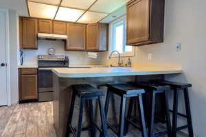 Kitchen with a kitchen breakfast bar, stainless steel electric stove, light countertops, under cabinet range hood, and brown cabinetry