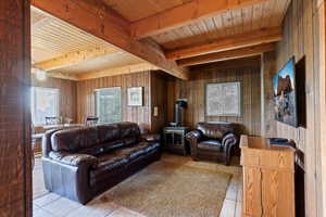 Living room featuring a wood stove, light tile patterned floors, wooden walls, and a wooden ceiling with exposed beams