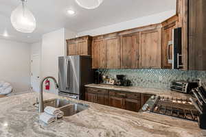 Kitchen with backsplash, light stone counters, hanging light fixtures, appliances with stainless steel finishes, and brown cabinets