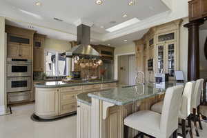 Kitchen with ornamental molding, glass insert cabinets, built in appliances, dark stone counters, and island exhaust hood