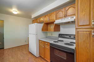 Kitchen with white appliances, under cabinet range hood, light wood-style flooring, light countertops, and brown cabinets