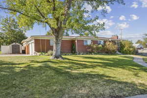 Ranch-style home featuring a front yard, brick siding, a storage unit, an attached garage, and a chimney