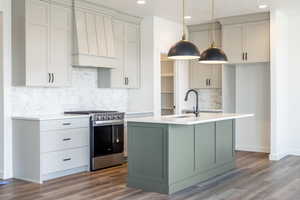 Kitchen with decorative backsplash, stainless steel range with gas stovetop, green cabinets, hanging light fixtures, and a center island with sink