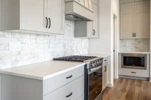 Kitchen featuring stainless steel appliances, custom exhaust hood, decorative backsplash, dark wood-type flooring, and light stone counters