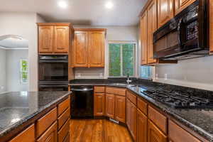 Kitchen featuring dark stone counters, dark wood-style floors, black appliances, brown cabinetry, and arched walkways
