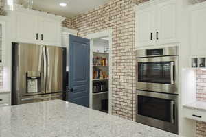 Kitchen featuring appliances with stainless steel finishes, white cabinets, light stone countertops, brick wall, and open shelves